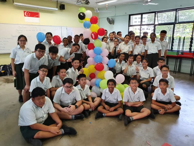 A large group of school students in uniforms posing in a classroom with a tall colorful balloon tower.